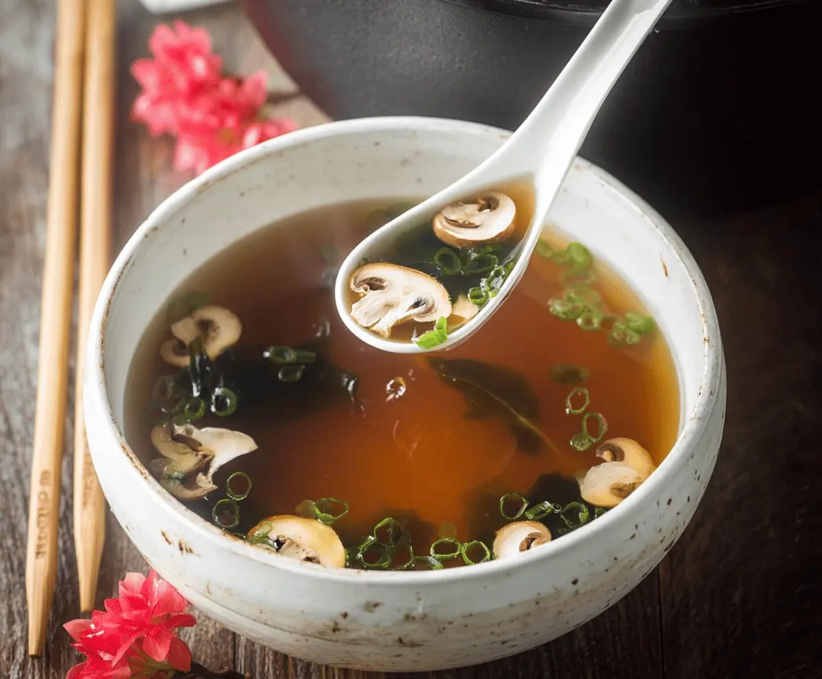 A bowl of traditional Japanese clear soup garnished with green onions, sliced mushrooms, and tofu cubes on a wooden table.